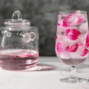 Crystal glass full of fresh beverage with pink rose petals and ice cubes place on table near jar with drink